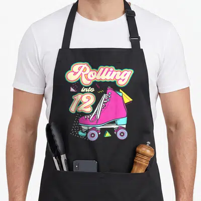 Tablier de cuisine noir avec illustration de roller rose et texte Rolling into 12 en lettres colorées style rétro.