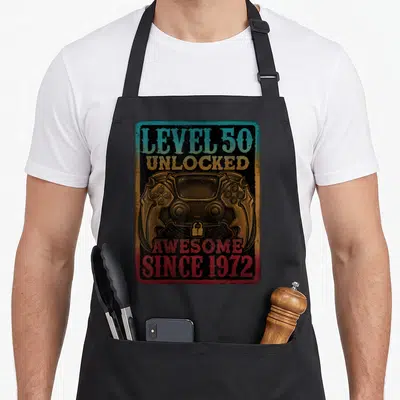 Tablier de cuisine noir avec illustration manette de jeu vidéo et texte Level 50 Unlocked Génial Depuis 1972 en style vintage.
