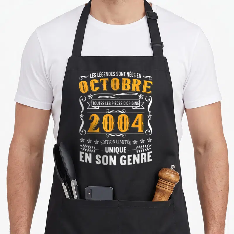 Tablier de cuisine noir avec typographie vintage orange et blanche citant les légendes nées en octobre 2004.