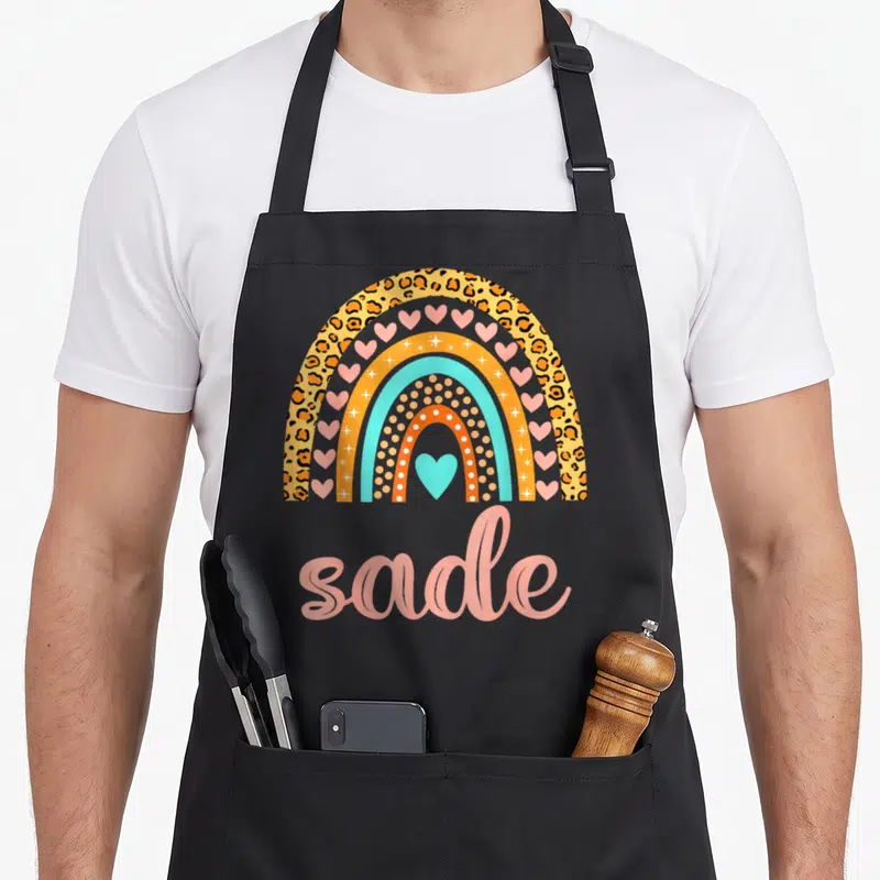 Tablier de cuisine noir avec arc-en-ciel coloré, motifs léopard, cœurs roses et le nom Sade en lettres cursives roses.
