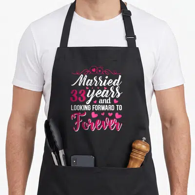 Tablier de cuisine noir avec l'inscription Married 33 years and Looking Forward to Forever en rose et blanc entouré de cœurs.