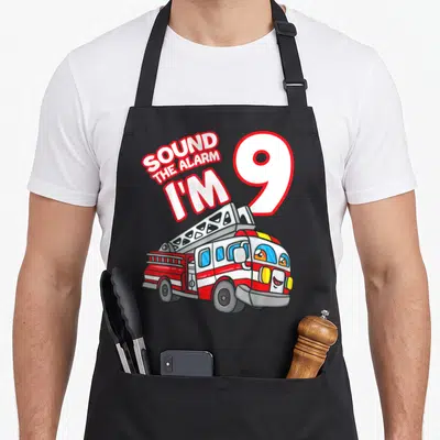Tablier de cuisine noir avec illustration de camion de pompier souriant et texte Sound The Alarm I'm 9 en rouge et blanc.
