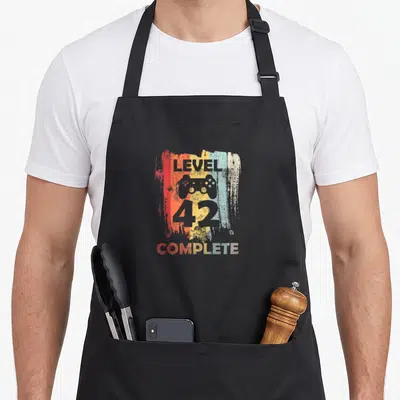 Tablier de cuisine noir avec motif Level 42 Complete et icône de manette de jeu vidéo sur fond texturé multicolore.