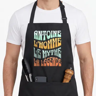 Tablier de cuisine noir avec typographie rétro multicolore affichant le prénom Antoine et des qualificatifs humoristiques.