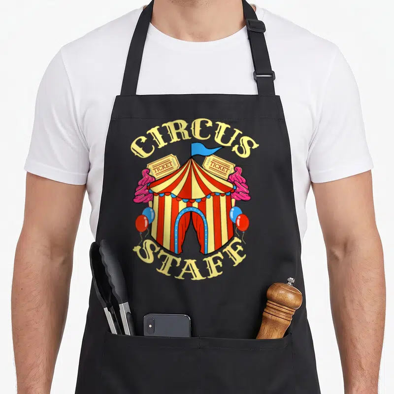Tablier de cuisine noir avec une tente de cirque rouge et jaune, des tickets et des ballons colorés.