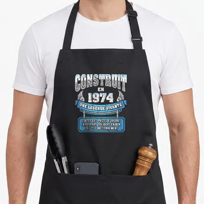 Tablier de cuisine noir avec inscription Construit en 1974 Une Légende Vivante et blason bleu humoristique.