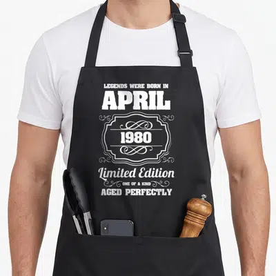 Tablier de cuisine noir avec inscription blanche « Legends were born in April 1980 Limited Edition Aged Perfectly » et cadre décoratif.