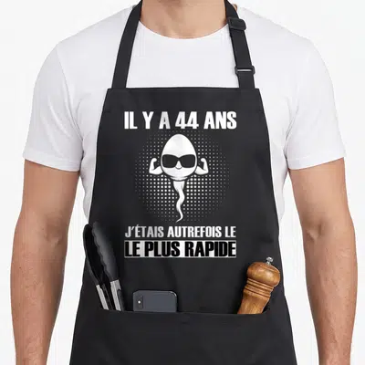 Tablier de cuisine noir avec illustration d'un spermatozoïde musclé portant des lunettes de soleil et texte blanc.