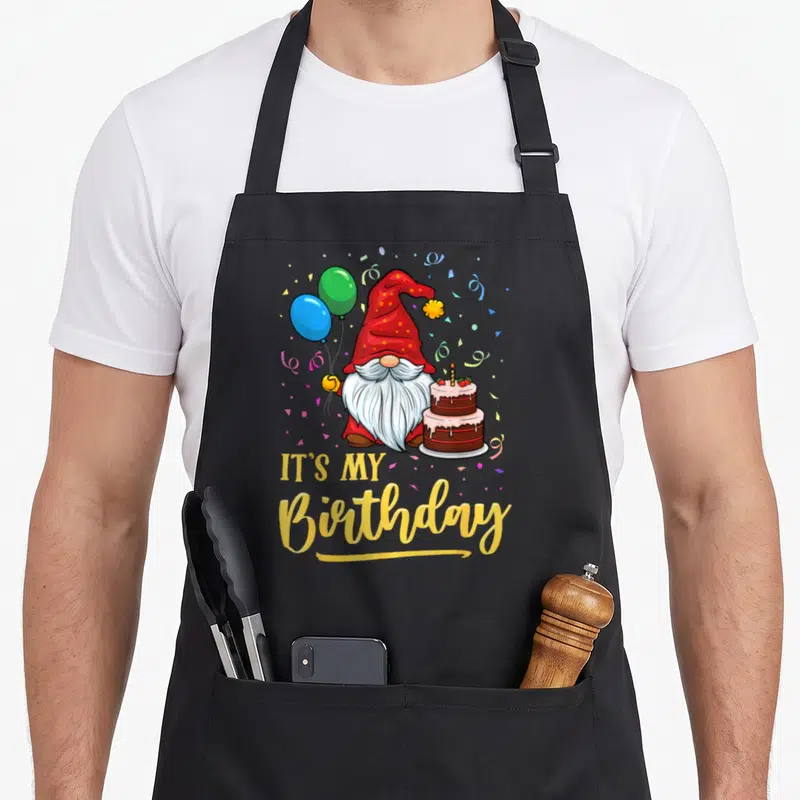 Tablier de cuisine noir avec un gnome rouge tenant des ballons et un gâteau d'anniversaire.