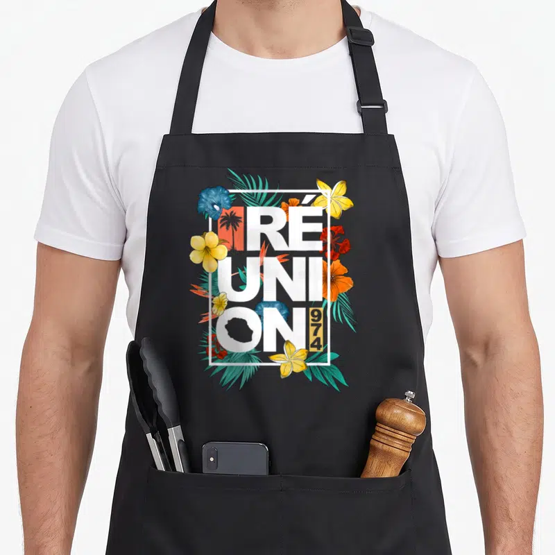 Tablier de cuisine noir avec inscription Réunion 974 entourée de fleurs tropicales jaunes, rouges et bleues ainsi que de feuilles vertes.