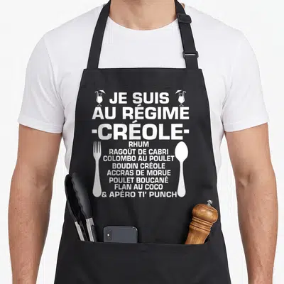Tablier de cuisine noir avec texte blanc listing les plats créoles et un verre à cocktail stylisé sur le côté.