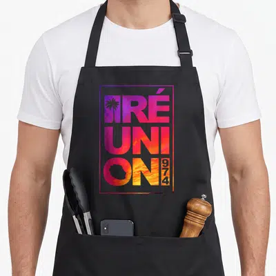 Tablier de cuisine noir avec typographie colorée Réunion 974 et silhouette de palmier sur fond nuageux.