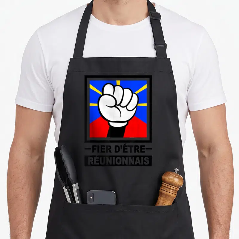 Tablier de cuisine noir avec illustration d'un poing levé blanc sur fond bleu rouge et jaune, suivi du texte FIER D'ÊTRE RÉUNIONNAIS.