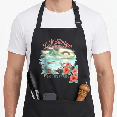 Tablier de cuisine noir avec illustration colorée d'un volcan, d'une pirogue, d'un colibri et de fleurs de hibiscus roses sur fond tropical.