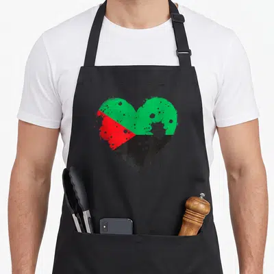 Tablier de cuisine noir avec un grand cœur stylisé aux couleurs vert rouge et noir évoquant le drapeau de la Martinique avec effet de peinture éclaboussée.