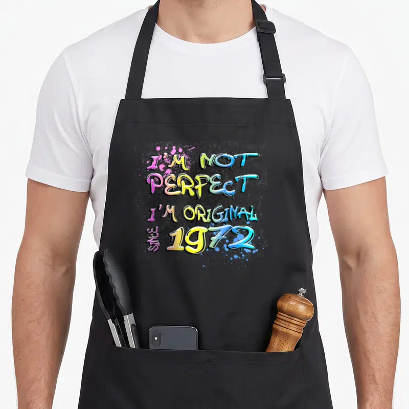 Tablier de cuisine noir avec inscription I'M NOT PERFECT I'M ORIGINAL SINCE 1972 en lettres graffiti multicolores.