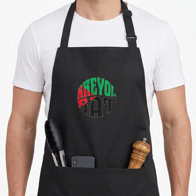 Tablier de cuisine noir avec l'inscription circulaire Kreyol Sat en vert rouge et noir.