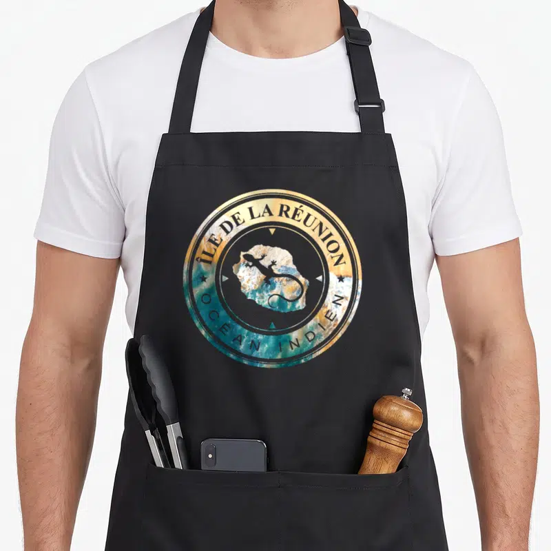Tablier de cuisine noir avec un logo circulaire illustrant l'île de la Réunion au milieu de l'Ocean Indien.