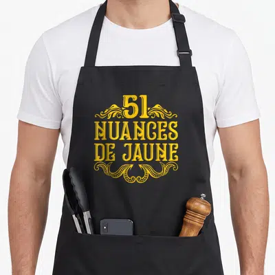 Tablier de cuisine noir avec inscription 51 Nuances de Jaune en lettres jaunes et motifs décoratifs vintage.