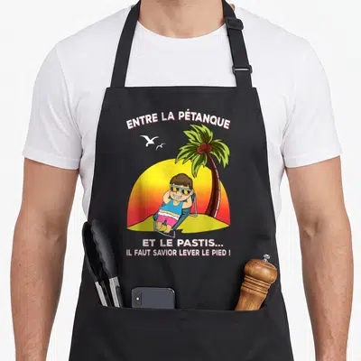 Tablier de cuisine noir avec illustration d'un homme en maillot de bain sous un palmier et le texte Entre la pétanque et le pastis.