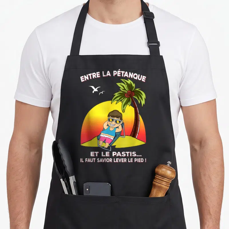 Tablier de cuisine noir avec illustration d'un homme en maillot de bain sous un palmier et le texte Entre la pétanque et le pastis.