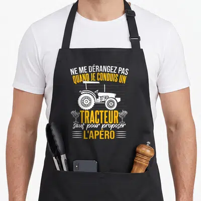 Tablier de cuisine noir avec illustration de tracteur blanc et texte jaune sur fond gris uni.