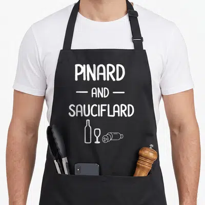 Tablier de cuisine noir avec le texte blanc Pinard and Sauciflard et des dessins de bouteille, verre et saucisson en bas.