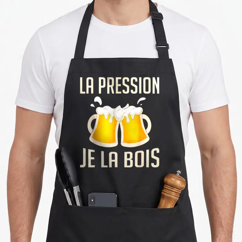 Tablier de cuisine noir avec l'inscription La pression je la bois au-dessus de deux verres de bière dorée qui se toisent.
