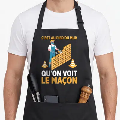 Tablier de cuisine noir avec illustration d'un maçon et le texte C'est au pied du mur qu'on voit le maçon.