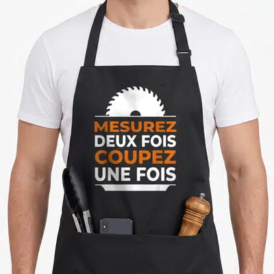 Tablier de cuisine noir avec une scie circulaire blanche et le texte Mesurez deux fois coupez une fois en orange et blanc.