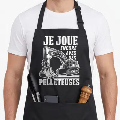 Tablier de cuisine noir avec illustration d'une excavatrice blanche et le texte Je joue encore avec des pelleteuses.