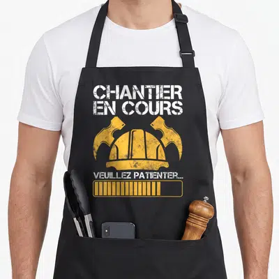 Tablier de cuisine noir avec casque de chantier jaune, deux marteaux croisés et texte blanc Chantier en cours.