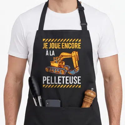 Tablier de cuisine noir avec illustration excavatrice orange et texte Je joue encore à la pelleteuse en blanc et jaune.