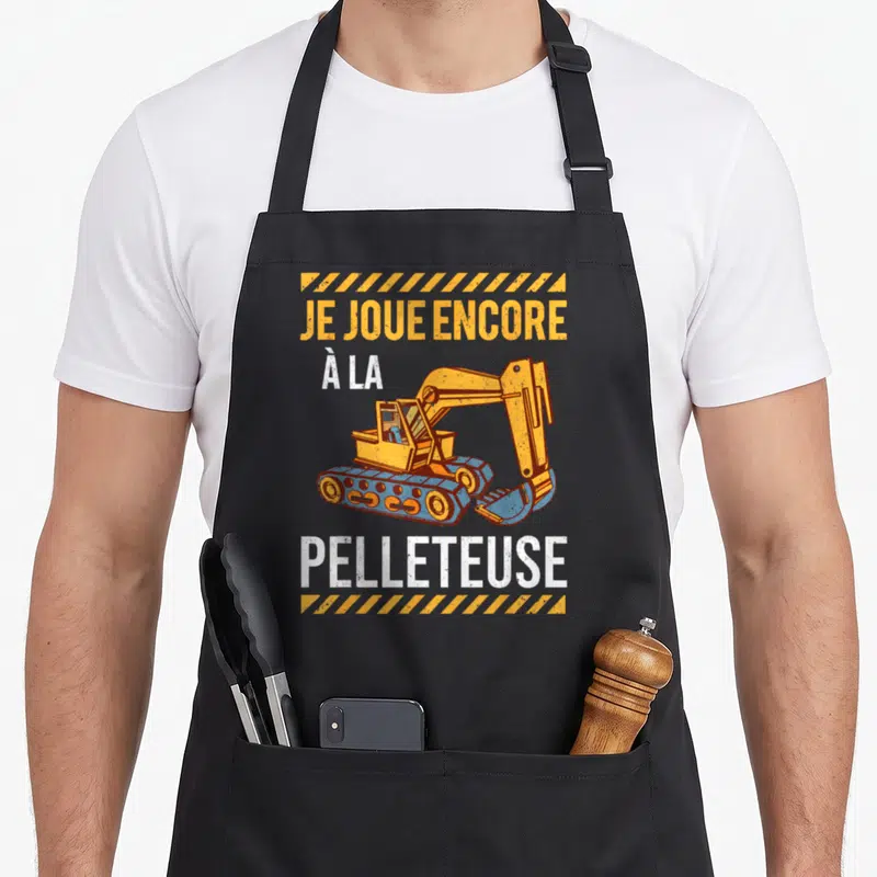 Tablier de cuisine noir avec illustration excavatrice orange et texte Je joue encore à la pelleteuse en blanc et jaune.