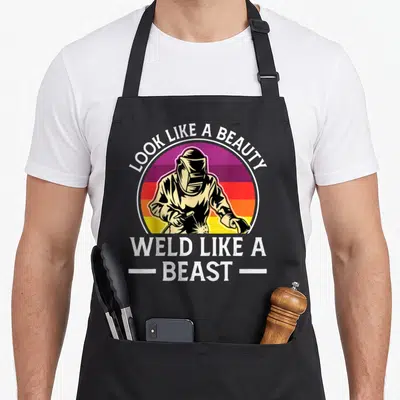 Tablier de cuisine noir avec illustration d'un soudeur et texte « Look like a beauty weld like a beast » sur fond coloré.