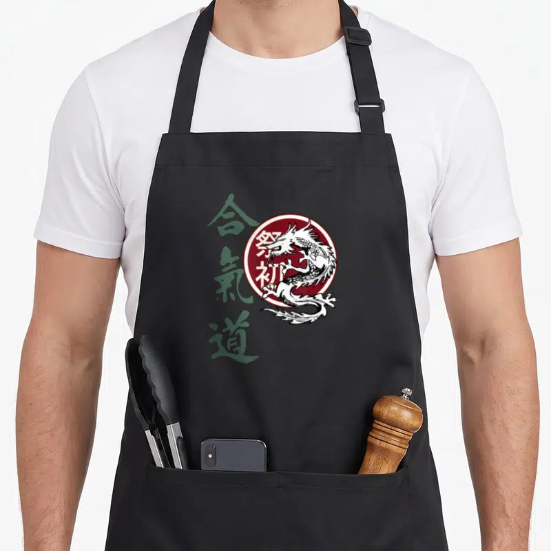 Tablier de cuisine noir avec un dragon blanc dans un cercle bordeaux et des caractères japonais verts sur le côté gauche.