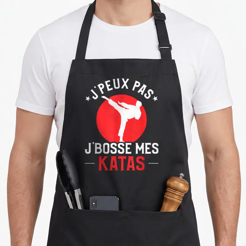 Tablier de cuisine noir avec silhouette blanche effectuant un coup de pied dans un cercle rouge et texte J'peux pas j'bosse mes katas