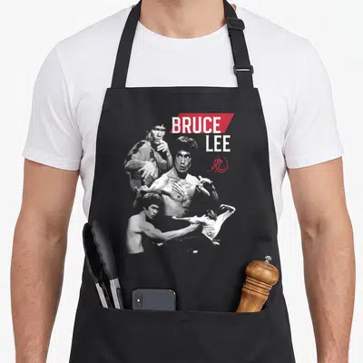 Tablier de cuisine noir avec un collage de Bruce Lee en position de combat et son nom imprimé en lettres blanches sur fond rouge.