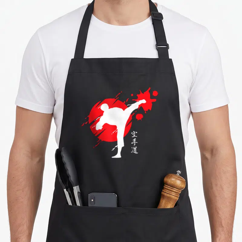 Tablier de cuisine noir avec silhouette blanche effectuant un coup de pied devant un cercle rouge et des caractères japonais.