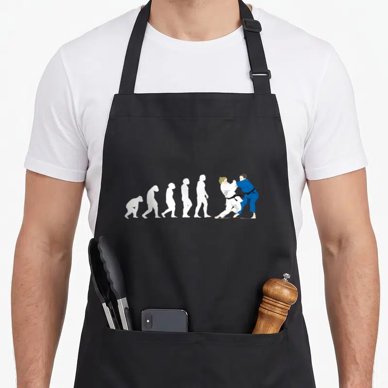 Tablier de cuisine noir avec une illustration montrant l'évolution humaine culminant par deux judokas en train de s'affronter sur le tatami.