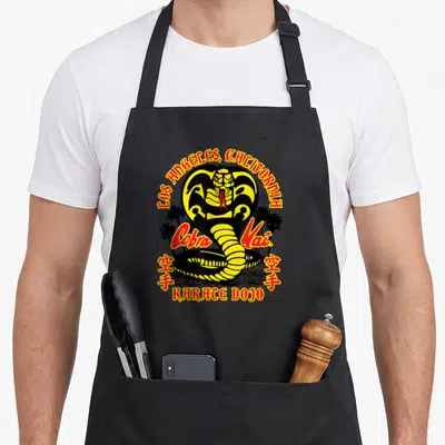 Tablier de cuisine noir avec un cobra jaune et noir, texte Cobra Kai, Karate Dojo et Los Angeles California entouré de palmiers.
