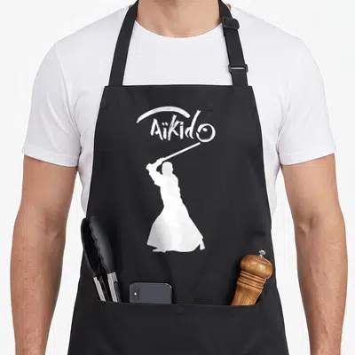 Tablier de cuisine noir avec silhouette blanche d'un pratiquant d'Aikido tenant un bâton sous le mot Aikido en style calligraphie.