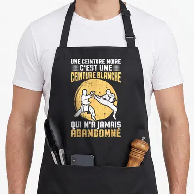 Tablier de cuisine noir avec deux silhouettes en combat et le texte Une ceinture noire c'est une ceinture blanche qui n'a jamais abandonné.