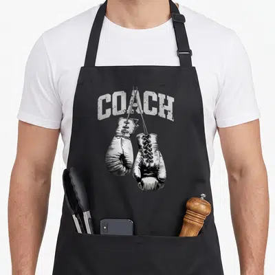 Tablier de cuisine noir avec illustration de gants de boxe et le texte COACH en lettres blanches usées.
