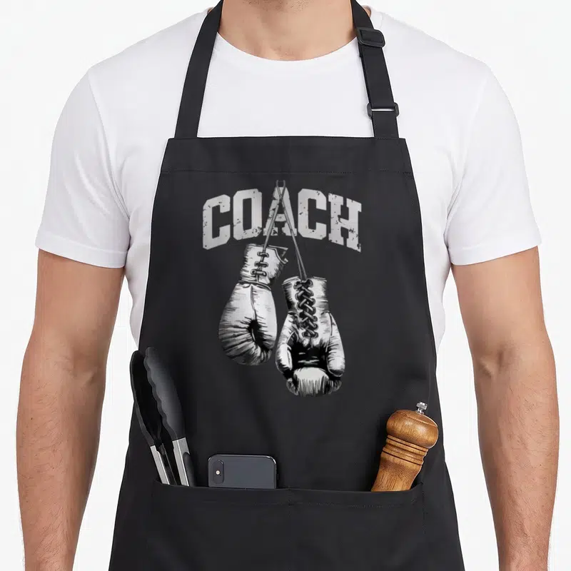 Tablier de cuisine noir avec illustration de gants de boxe et le texte COACH en lettres blanches usées.