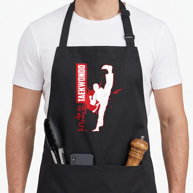 Tablier de cuisine noir avec silhouette blanche de Taekwondo, texte rouge vertical et éclaboussures artistiques sur fond uni.