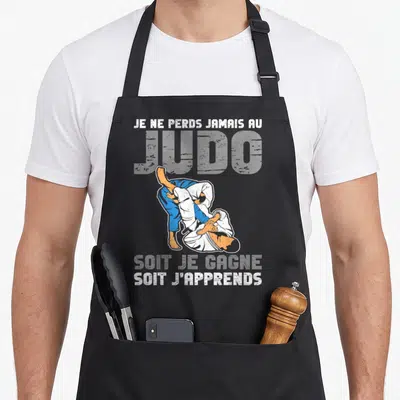 Tablier de cuisine noir avec une illustration de deux judokas en action et un texte humoristique sur la victoire et l'apprentissage.