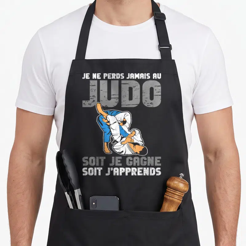 Tablier de cuisine noir avec une illustration de deux judokas en action et un texte humoristique sur la victoire et l'apprentissage.