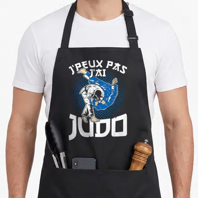 Tablier de cuisine noir avec illustration d'un judoka en action et le texte J'peux pas j'ai judo imprimé en blanc et bleu.