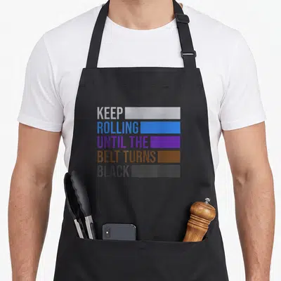 Tablier de cuisine noir avec inscription Keep Rolling Until the Belt Turns Black et bandes colorées représentant les ceintures.
